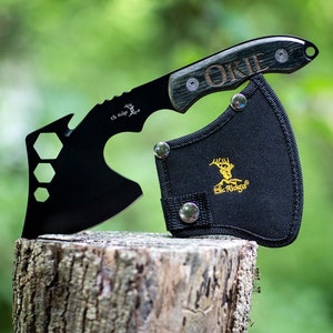 May include: A black Elk Ridge hatchet with a wooden handle and the word "OKIE" etched on it. The hatchet has a bottle opener and hexagonal cutouts. It rests on a tree stump next to a black sheath with the Elk Ridge logo.