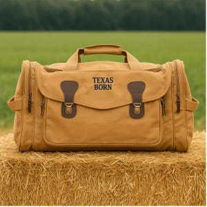 May include: A tan canvas duffel bag with brown leather accents and the text "TEXAS BORN" in black. The bag has a top handle, side pockets, and a front flap with buckles. It is sitting on a bale of hay.