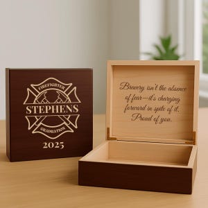May include: A wooden box with a hinged lid, featuring a dark brown exterior and a lighter wood interior. The front of the box is engraved with "FIREFIGHTER," "STEPHENS," "GRADUATION," and the year "2025." The open box reveals an inscription.