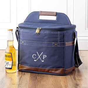 May include: A navy blue canvas cooler bag with brown leather accents. The cooler bag has a top handle, a shoulder strap, and a zipper closure. The bag is personalized with the initials "CXP" and a pair of crossed golf clubs.