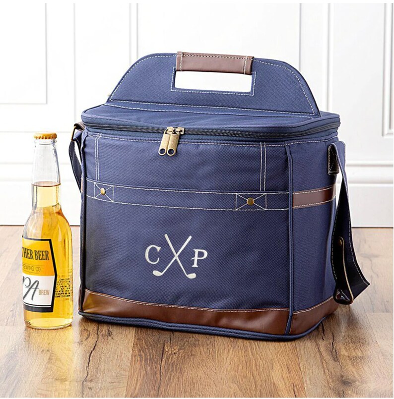 May include: A navy blue canvas cooler bag with brown leather accents. The cooler bag has a top handle, a shoulder strap, and a zipper closure. The bag is personalized with the initials "CXP" and a pair of crossed golf clubs.