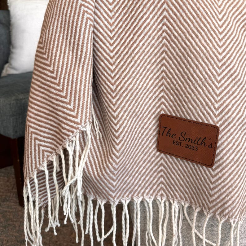 K&ouml;nnte beinhalten: Eine braune und wei&szlig;e Chevron-Muster-Decke mit Fransenrand. Ein braunes Leder-Patch mit dem Text "The Smith's Est. 2023" ist auf die Decke gen&auml;ht.