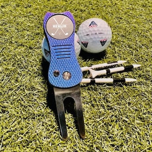 May include: A blue golf ball marker with the text "KEKLIK" and two crossed golf clubs. The marker is attached to a silver golf ball retriever with a blue and silver finish. The marker and retriever are on a green artificial grass surface with two white golf balls and three white and black golf tees.