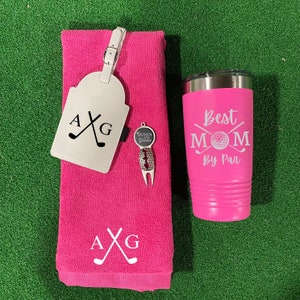 May include: A pink golf towel with the initials "AXG" embroidered on it, a white leather luggage tag with the same initials and crossed golf clubs, a silver golf divot tool with a "Queen of the Green" charm, and a pink tumbler with the text "Best Mom By Par" and a golf ball graphic.