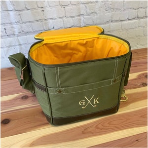May include: A green canvas cooler bag with a brown leather trim and a yellow interior. The bag has a zipper closure and a shoulder strap. The bag is monogrammed with the initials "G X K" and a golf club design.