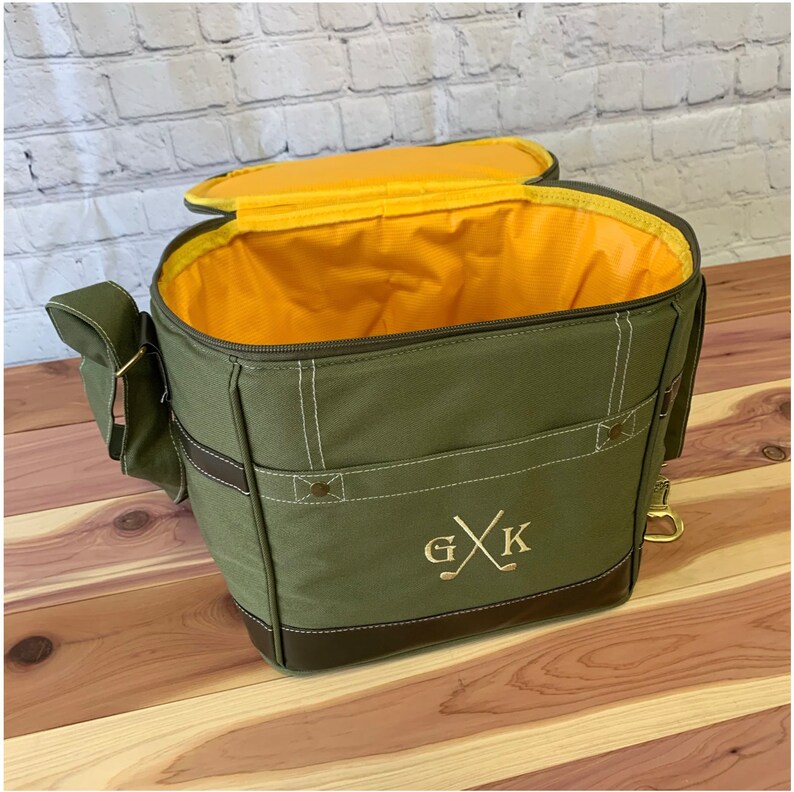 May include: A green canvas cooler bag with a brown leather trim and a yellow interior. The bag has a zipper closure and a shoulder strap. The bag is monogrammed with the initials "G X K" and a golf club design.