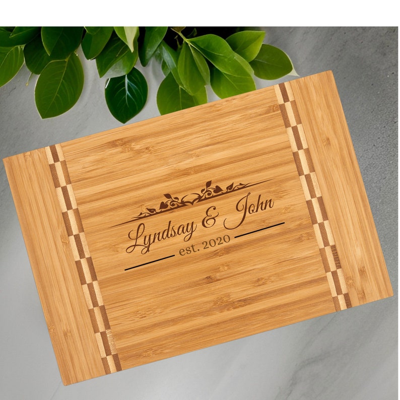 Może przedstawiać: Prostokątna deska do krojenia z bambusa z wygrawerowanym tekstem "Lyndsay & John est. 2020". Deska ma dekoracyjne wzory w szachownicę po bokach i kwiatowy wz&oacute;r nad imionami. Deska jest jasnobrązowa.