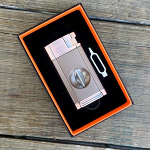 May include: A brown and silver cigar cutter and lighter combo in a black box with an orange border. The cutter has a round blade and the lighter has a silver finish.