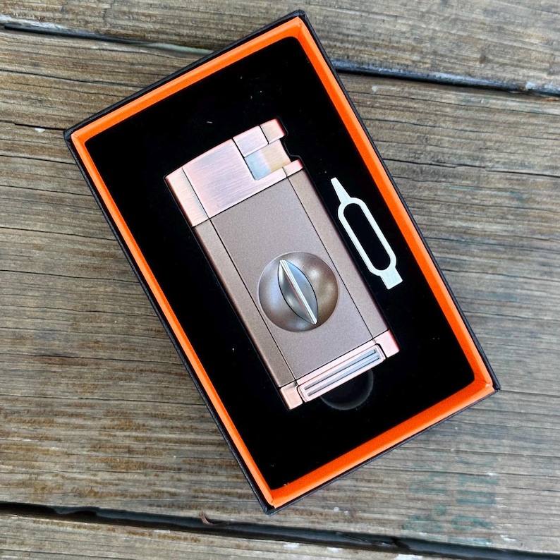 May include: A brown and silver cigar cutter and lighter combo in a black box with an orange border. The cutter has a round blade and the lighter has a silver finish.