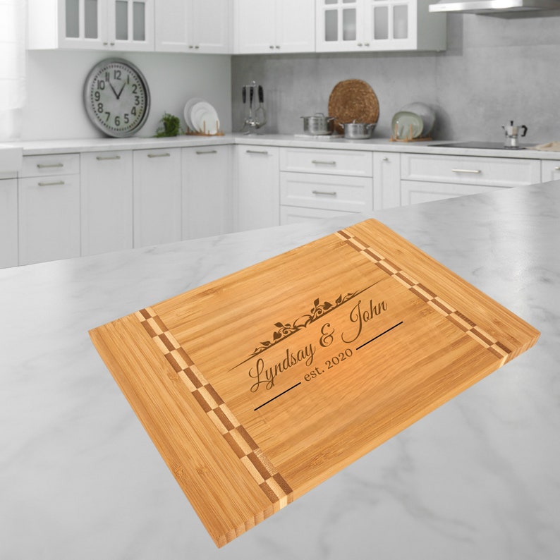Może przedstawiać: Prostokątna deska do krojenia z bambusa z ozdobną obw&oacute;dką i wygrawerowanym tekstem. Tekst brzmi "Lindsay & John est. 2020" z kwiatowymi akcentami. Deska jest jasnobrązowa z ciemnobrązowymi akcentami.