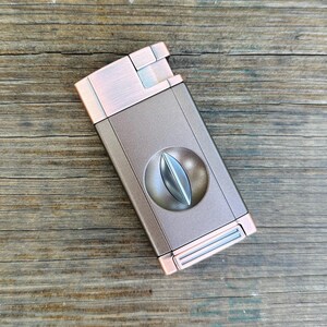 May include: A copper and silver cigar cutter with a guillotine blade. The cutter has a brushed metal finish and a rounded shape.