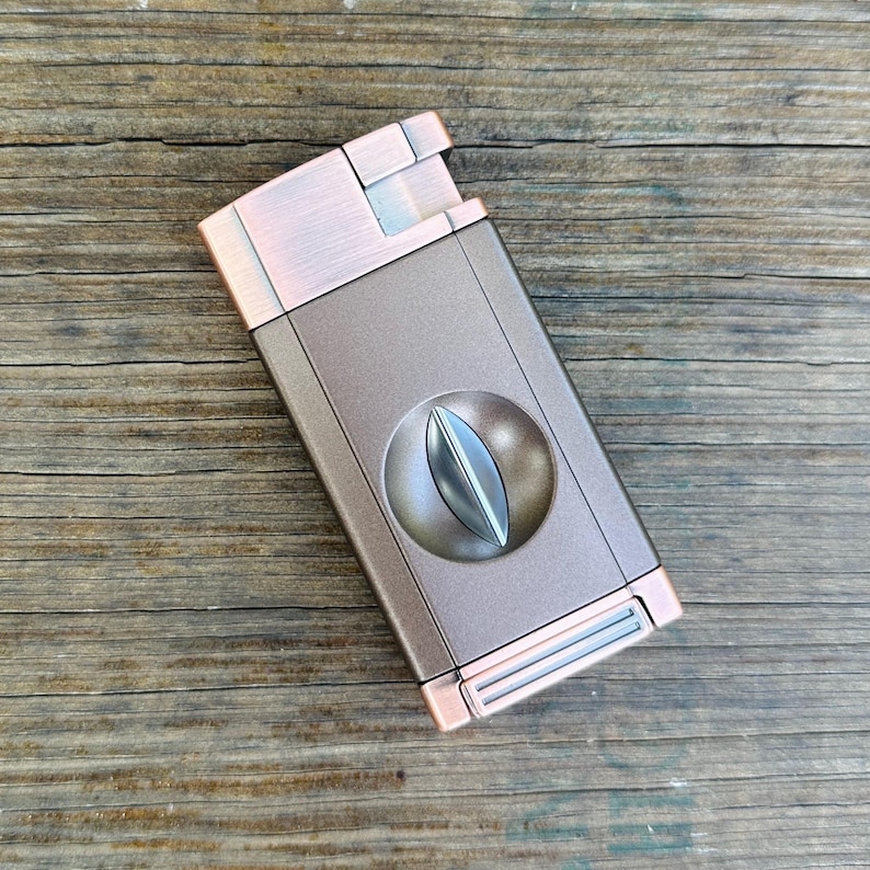 May include: A copper and silver cigar cutter with a guillotine blade. The cutter has a brushed metal finish and a rounded shape.