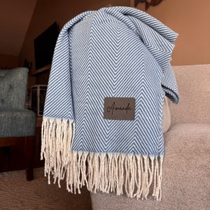 K&ouml;nnte beinhalten: Eine blaue und wei&szlig;e Chevron-Muster-Wolldecke mit einem braunen Lederanh&auml;nger, auf dem "Amanda" steht, und wei&szlig;en Fransen am unteren Rand.