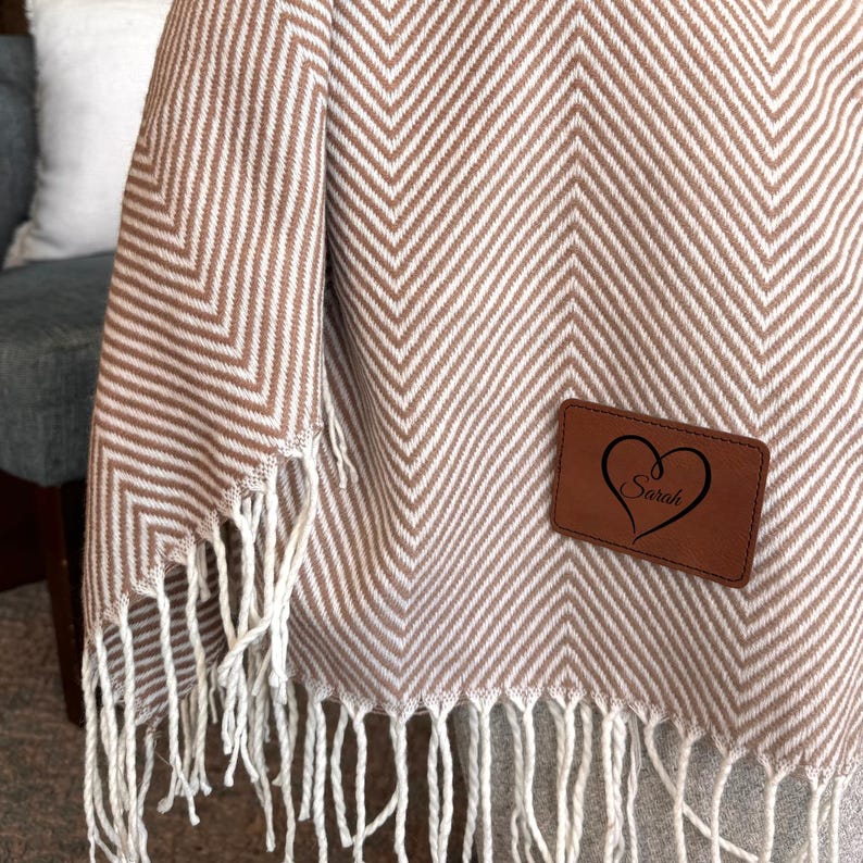 K&ouml;nnte beinhalten: Eine braune und wei&szlig;e Chevron-Muster-Decke mit Fransenrand. Ein braunes Leder-Herz-Etikett mit dem Namen "Sarah" ist an der Decke befestigt.