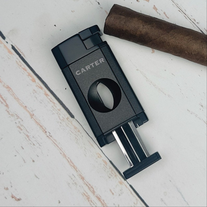 May include: Black cigar cutter with the word "CARTER" printed on the side. The cutter has a circular blade and a sliding mechanism to adjust the size of the cut.