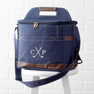 May include: A navy blue canvas cooler bag with a brown leather handle and trim. The bag has a zippered top and a shoulder strap. The bag is monogrammed with the initials "CXP" and a pair of crossed golf clubs.