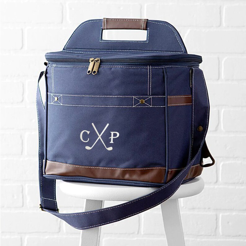 May include: A navy blue canvas cooler bag with a brown leather handle and trim. The bag has a zippered top and a shoulder strap. The bag is monogrammed with the initials "CXP" and a pair of crossed golf clubs.