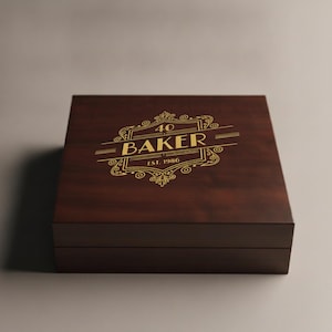 May include: A square dark brown wooden box with a hinged lid. The top of the box features a gold-colored decorative emblem with the text 40 BAKER EST. 1986 centered within an ornate frame consisting of lines and swirling flourishes.