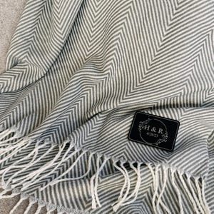 K&ouml;nnte beinhalten: Graue und wei&szlig;e Chevron-Muster-Decke mit wei&szlig;en Fransen. Die Decke hat ein kleines schwarzes Etikett mit dem Text "H&R 4/28/23".