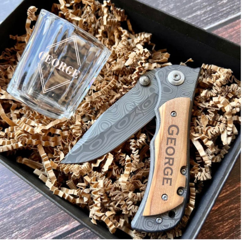 May include: A gift set with a personalized pocket knife and a shot glass. The knife has a wooden handle with the name "GEORGE" engraved on it. The shot glass is clear glass with the name "GEORGE" engraved on it.