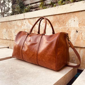 May include: A brown leather duffel bag with a top handle and a shoulder strap. The bag has a zipper closure and a front pocket.
