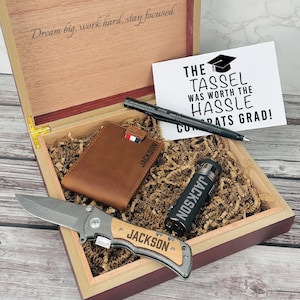 May include: A wooden gift box with a brown leather wallet, a black pen, a black lighter, a pocket knife, and a card that reads "The tassel was worth the hassle Congrats Grad!"