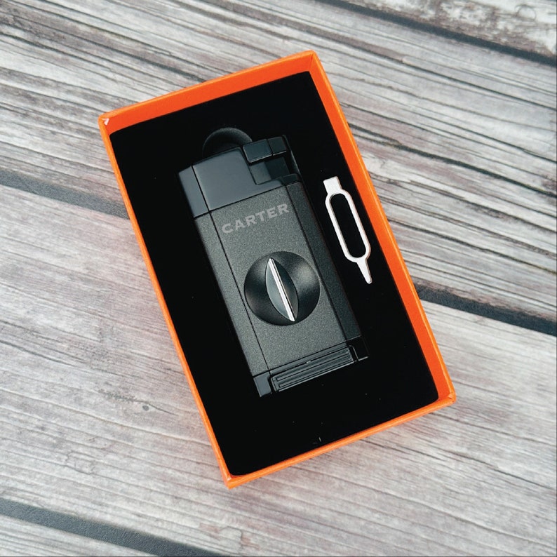 May include: A black cigar cutter with the word "CARTER" printed on it, in an orange box with a small silver tool.