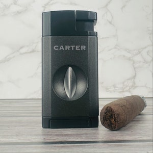 May include: A black cigar cutter with the word "CARTER" printed on the front. The cutter has a silver blade and is next to a brown cigar.