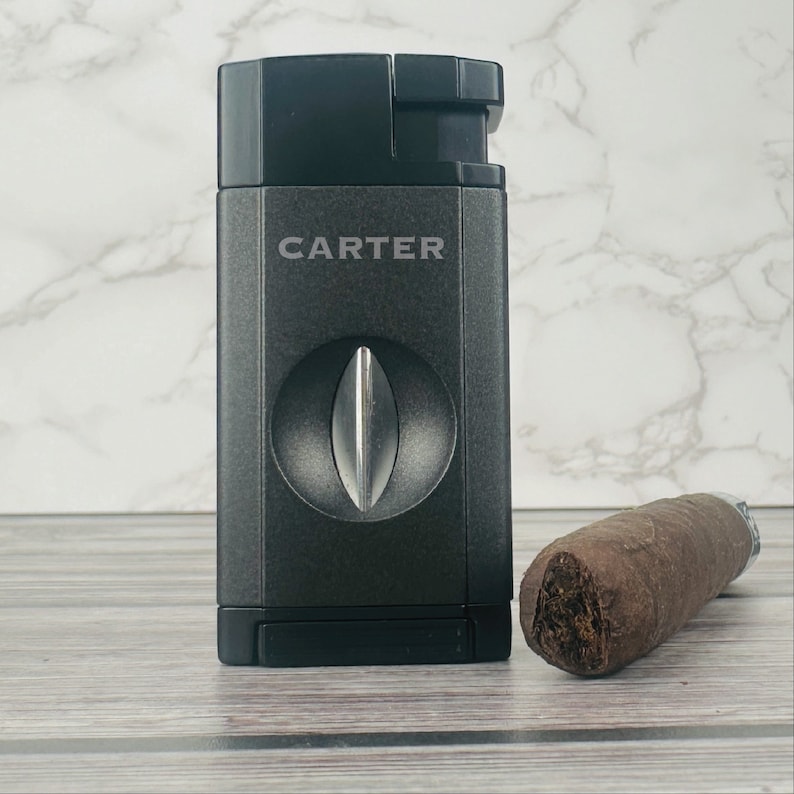 May include: A black cigar cutter with the word "CARTER" printed on the front. The cutter has a silver blade and is next to a brown cigar.