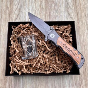 May include: A black and wood folding pocket knife with a personalized engraved handle that reads "GEORGE". The knife is in a black gift box with brown shredded paper and a small clear shot glass with the same engraved name.