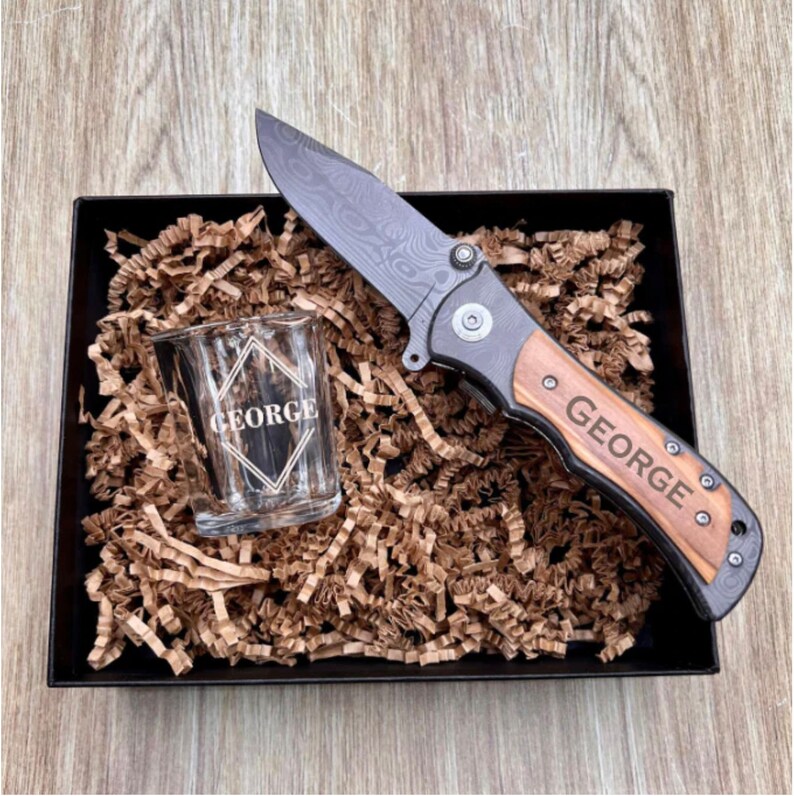 May include: A black and wood folding pocket knife with a personalized engraved handle that reads "GEORGE". The knife is in a black gift box with brown shredded paper and a small clear shot glass with the same engraved name.