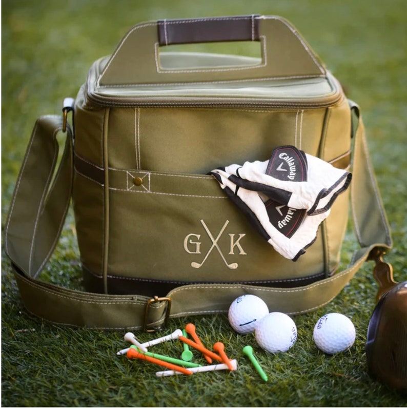 May include: A green canvas cooler bag with a brown leather handle and a brown leather trim. The bag has a zipper closure and a large pocket on the front. The bag is monogrammed with the initials "G X K". The bag is sitting on a green grassy surface with golf balls and tees.