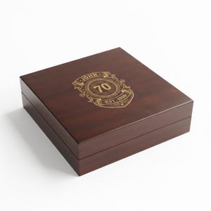 May include: A square dark brown wooden box with a hinged lid. The top of the lid features a gold-colored engraved emblem with a circular border, the name JOHN, the number 70, and the text EST. 1956, all arranged within a decorative ribbon banner.
