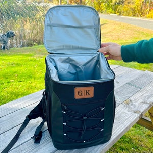 Pode incluir: Mochila cooler preta isolada com um remendo de couro marrom que diz "G|K". O cooler tem um bolso frontal com z&iacute;per e um fecho de cord&atilde;o. A mochila est&aacute; aberta e o interior &eacute; forrado com isolamento prateado.