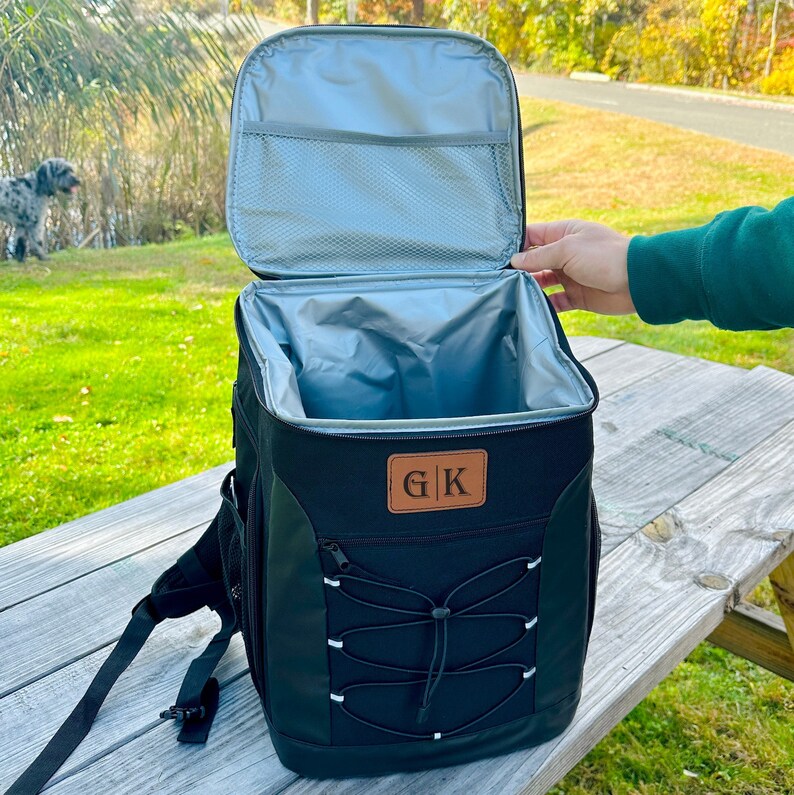 Pode incluir: Mochila cooler preta isolada com um remendo de couro marrom que diz "G|K". O cooler tem um bolso frontal com z&iacute;per e um fecho de cord&atilde;o. A mochila est&aacute; aberta e o interior &eacute; forrado com isolamento prateado.