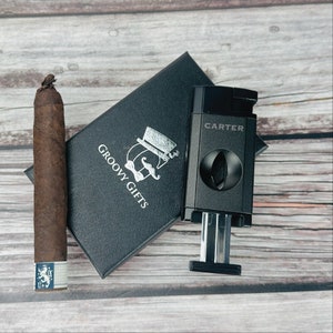 May include: A black cigar cutter with the word "CARTER" printed on it. The cutter is open and ready to cut a cigar. The cutter is sitting on a black box with the words "GROOVY GIFTS" printed on it. A brown cigar is sitting next to the box.