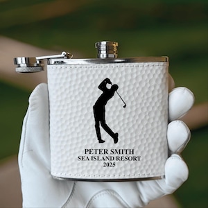 May include: A white, textured flask with a silver cap and hinge. A black silhouette of a golfer swinging a club is on the front, with the text "PETER SMITH SEA ISLAND RESORT 2025" below. The flask is held in a white glove.
