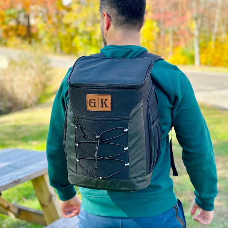 Pode incluir: Uma mochila cooler preta com um remendo de couro marrom que diz "G|K". A mochila tem um fecho de cord&atilde;o e al&ccedil;as ajust&aacute;veis.