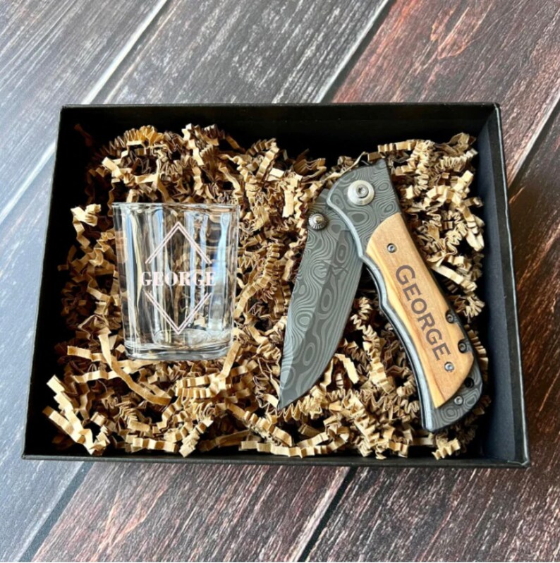 May include: A gift set in a black box with brown paper filler. The set includes a small shot glass with the name "George" engraved on it and a pocket knife with a wooden handle and the name "George" engraved on it.