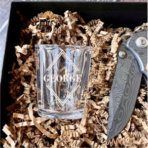 May include: A personalized shot glass with the name "GEORGE" etched in a geometric design. The glass is sitting in brown packing peanuts next to a black pocket knife.