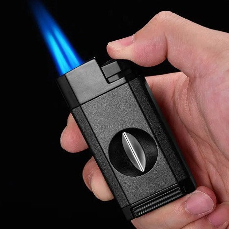 May include: A black cigar cutter and lighter with a blue flame. The cutter has a round blade and is held in a hand.