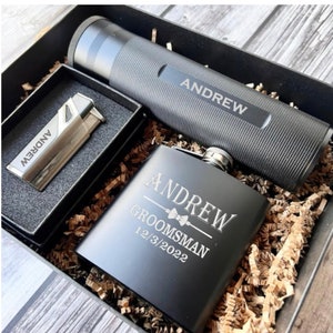 May include: A black gift box with a black flask, a silver lighter, and a black tube. The flask, lighter, and tube are engraved with the name "Andrew" and the text "Groomsman 12/3/2022".