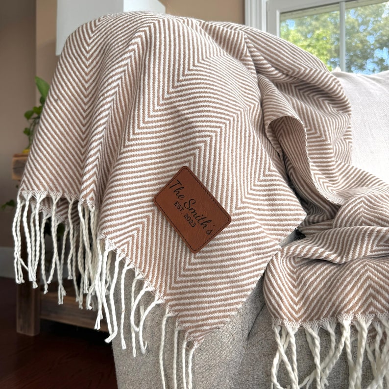 K&ouml;nnte beinhalten: Eine braune und wei&szlig;e Chevron-Muster-Wurfdecke mit Fransen. Die Decke hat ein braunes Leder-Etikett mit dem Text "The Smith's Est. 2023".