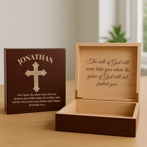 May include: A wooden keepsake box with a cross design and the name "JONATHAN" engraved on the lid. The box is open, revealing a light-colored interior with an inspirational quote. The exterior is a dark brown color, and the box is rectangular.