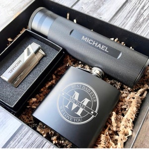 May include: A black gift box with a black flask, a silver lighter, and a black tube. The flask is engraved with the text "Best Dad Ever" and a monogram "M" with the name "Michael". The lighter and tube are also engraved with the name "Michael".