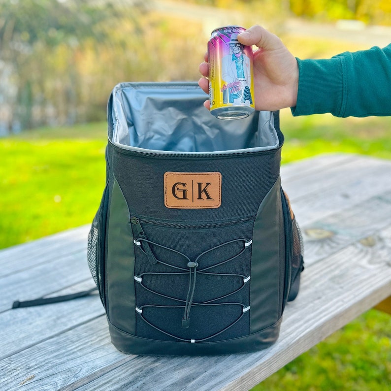 Pode incluir: Mochila t&eacute;rmica preta isolada com um remendo de couro marrom que diz "G|K". Uma m&atilde;o est&aacute; colocando uma lata de cerveja na geladeira.
