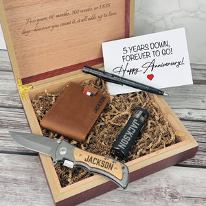 May include: A wooden gift box with a brown leather wallet, a black lighter, a black pen, and a pocket knife. The box is filled with brown shredded paper. The lid of the box has a personalized inscription that reads "Five years, 60 months, 260 weeks, or 1,825 days - however you count it, it all adds up to love." A white card with the text "5 YEARS DOWN, FOREVER TO GO! Happy Anniversary!" is inside the box.