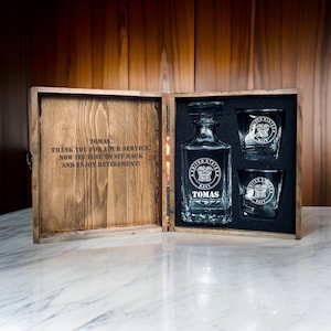 May include: A wooden gift box with a personalized whiskey decanter and two glasses. The decanter and glasses are etched with the United States Navy emblem and the name "TOMAS". The box is open, revealing the contents and a message.