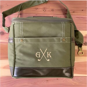 May include: A green canvas cooler bag with a brown leather trim and a zipper closure. The bag has a monogrammed "G X K" with crossed golf clubs embroidered on the front.