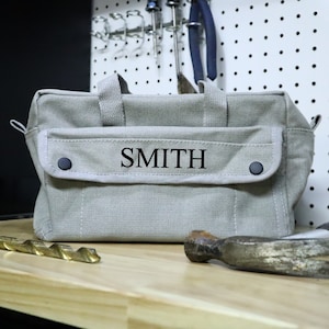 May include: A gray canvas tool bag with a black zipper and a black flap with the name "SMITH" embroidered on it.